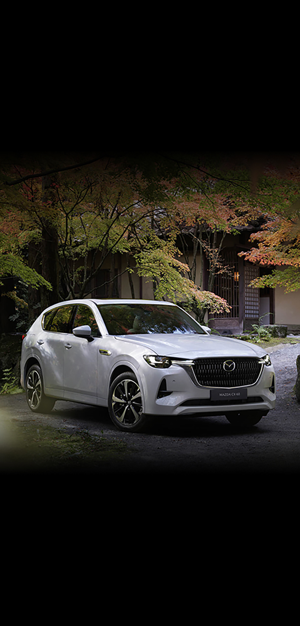 Il nuovo SUV Ibrido Plug-In Mazda CX-60 mostrato dalla parte anteriore parcheggiato all’esterno in un prato pieno di alberi.