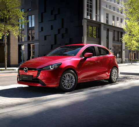 A red Mazda2 parked in front of a modern city building shown from the front and side.