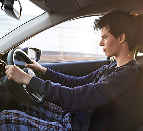 A young man behind the wheel of the Mazda2, driving down the road.