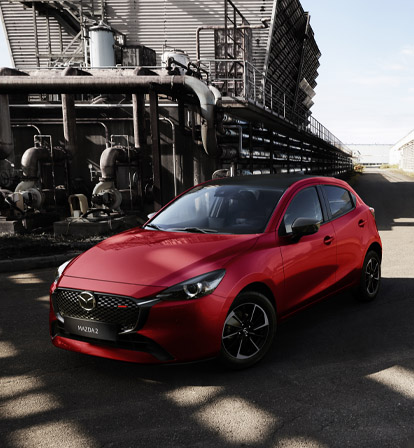 Una Mazda2 rossa ripresa frontalmente in un paesaggio industriale.