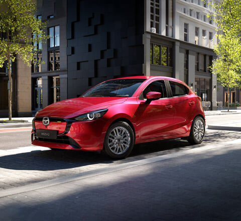 Una Mazda2 rossa parcheggiata di fronte a un edificio cittadino moderno e ripresa dalla parte anteriore e laterale.