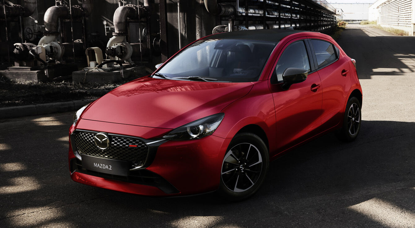 Una Mazda2 rossa ripresa frontalmente in un paesaggio industriale.