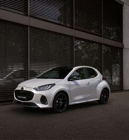 Mazda2 Hybrid v bílé barvě zaparkovaná před budovou.