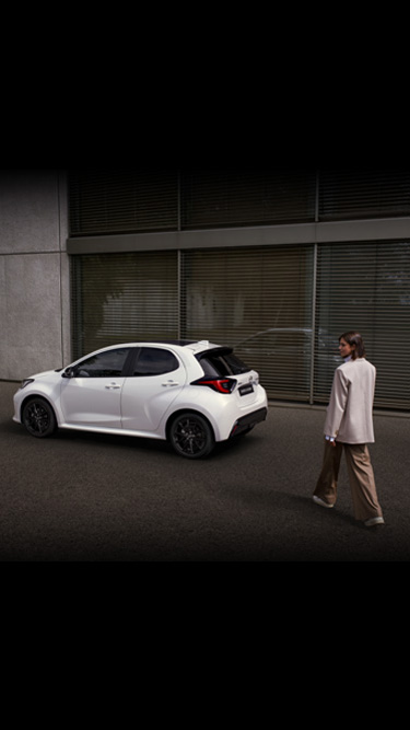 A white Mazda2 Hybrid seen the side parked in front of a building.