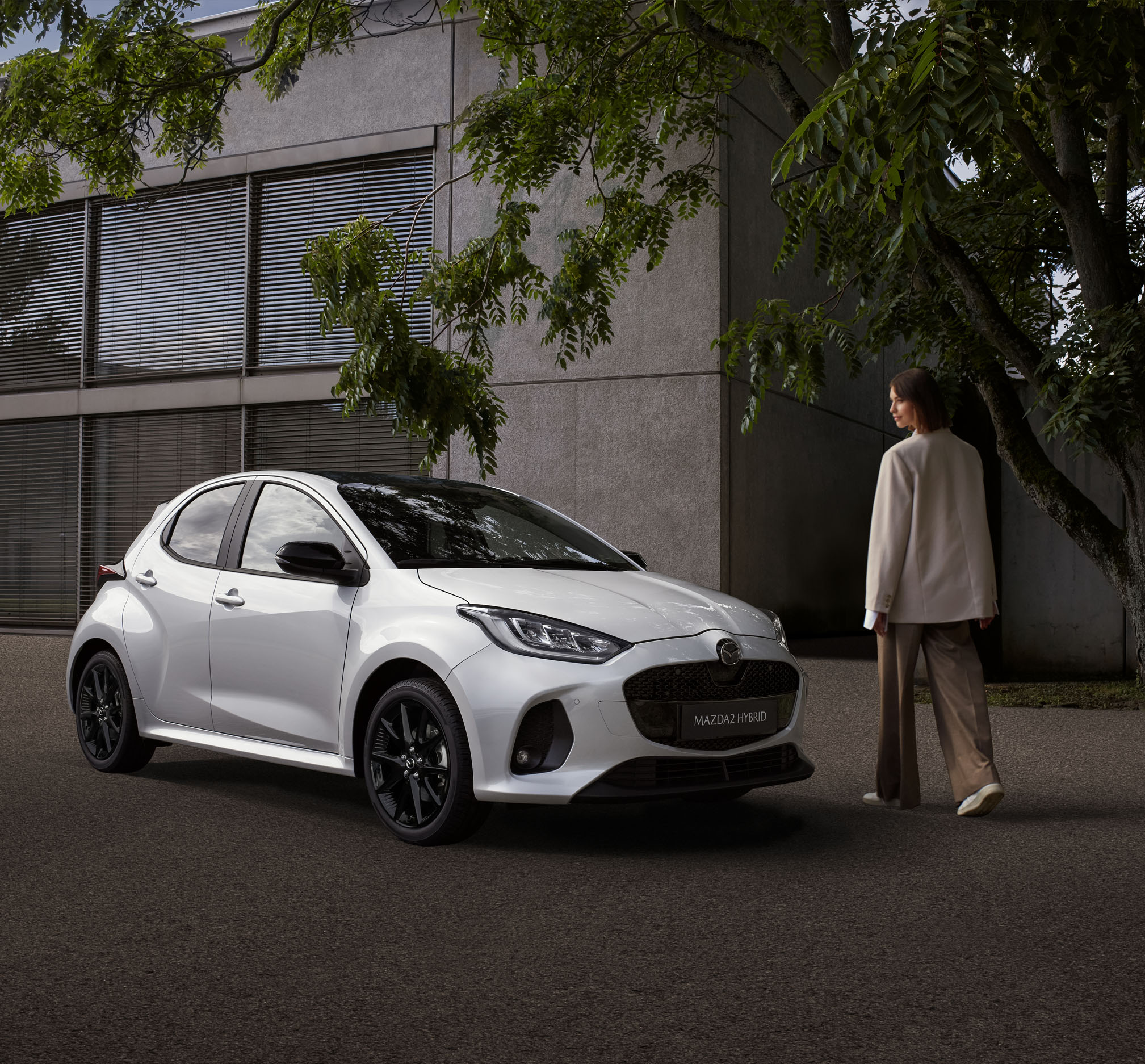 A woman walking next to the Mazda2 Hybrid parked in front a building.