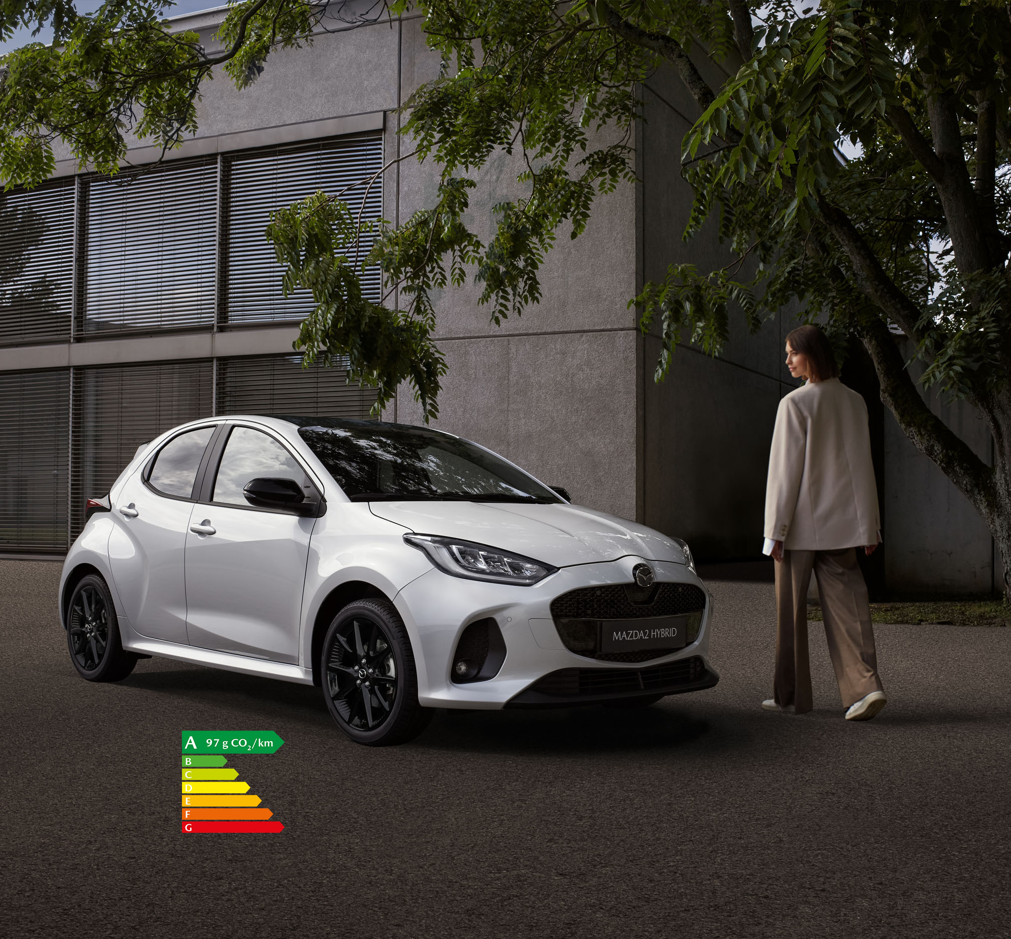 Femme marchant à côté d’une Mazda2 Hybrid stationnée devant un bâtiment.