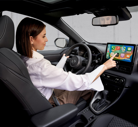 Donna che utilizza un pannello touchscreen mentre guida Mazda2 Hybrid.