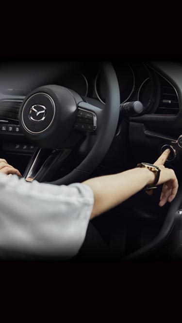 Μια γυναίκα στο εσωτερικό του Mazda3 αγγίζει το τιμόνι και πιέζει το κουμπί εκκίνησης.