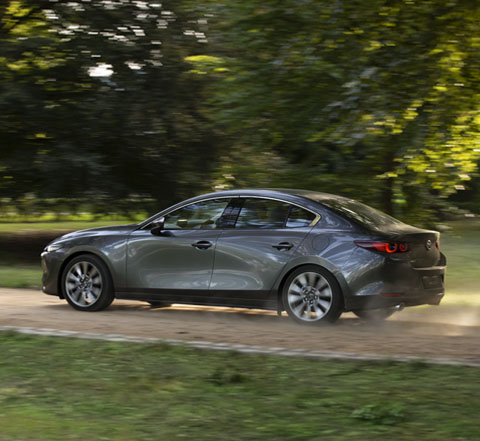 A grey Madza 3 Saloon on a country road shown from the side.