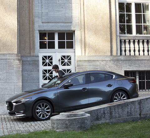 A grey Madza 3 Saloon parked in a sloped driveway with a man walking next to it.