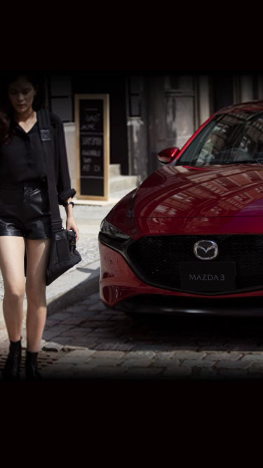 Woman with black dress holding a camera walks next to the left side of a red Madza3.