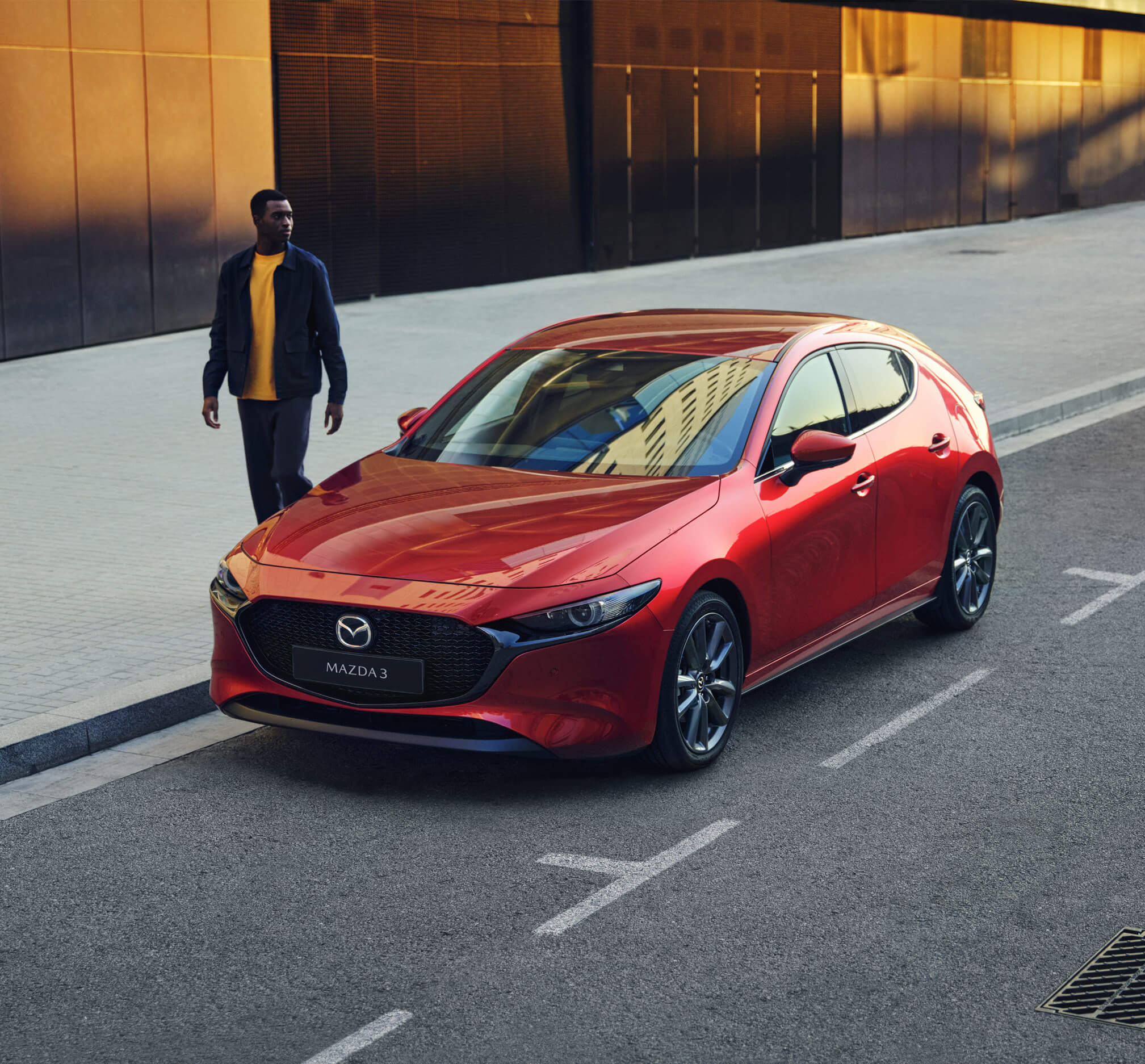 Un hombre con camiseta amarilla pasando junto a un Mazda3 5 puertas rojo.