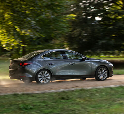 Šedá Mazda3 Sedan na venkovské cestě při pohledu z boku.