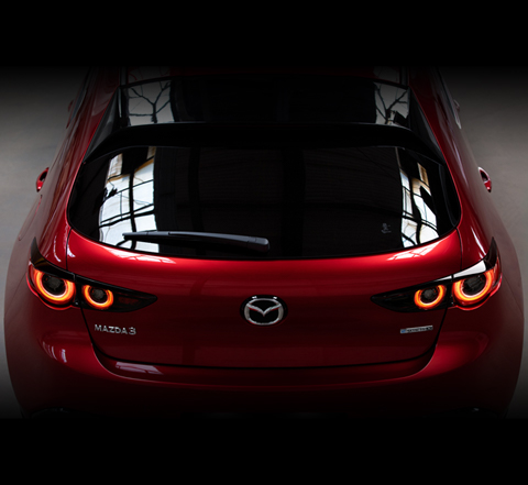 Das Heck eines roten Mazda3 Hatchback mit eingeschaltetem Licht.