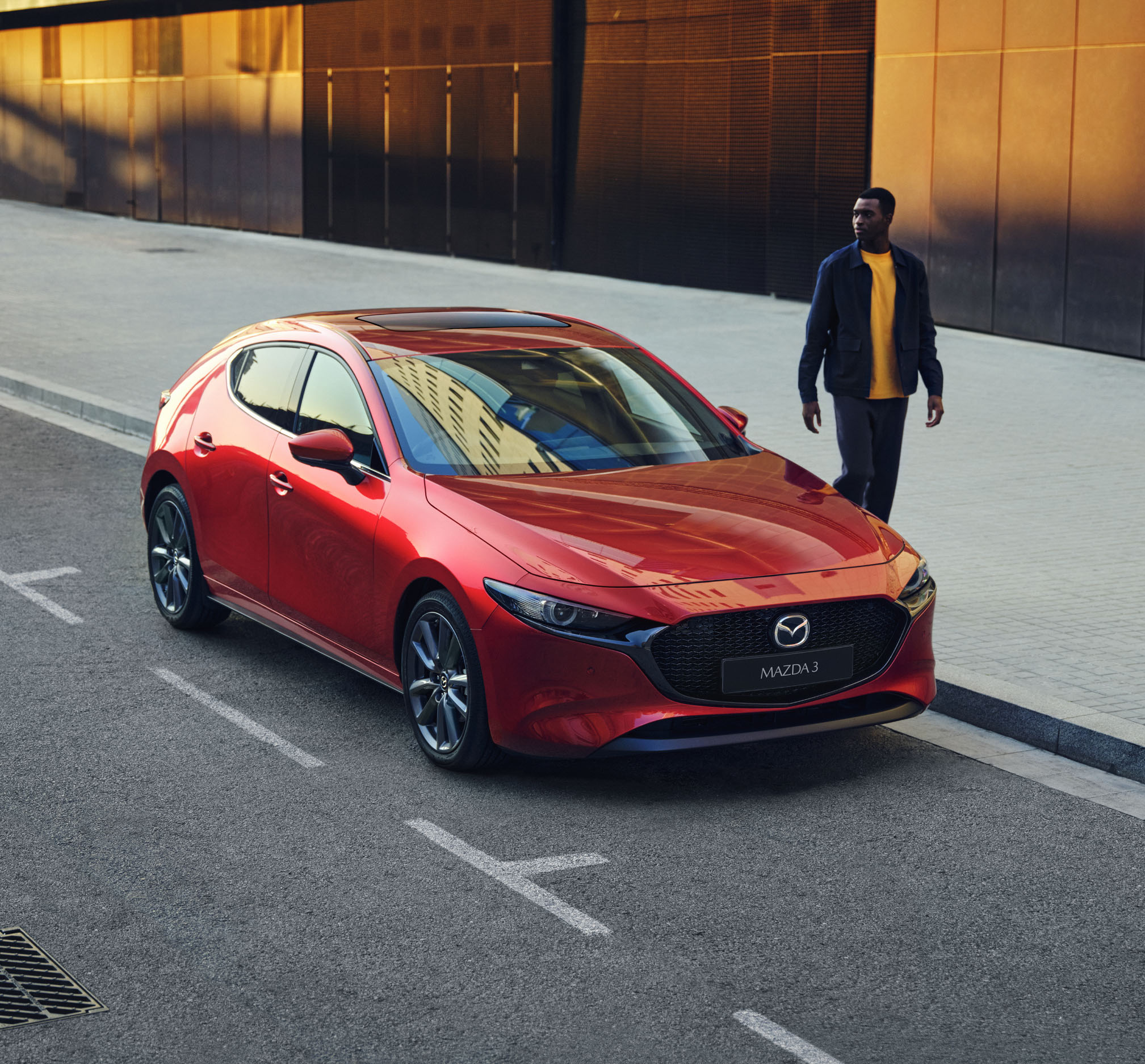 A man with yellow t-shirt walking next to a red Madza 3 hatchback.