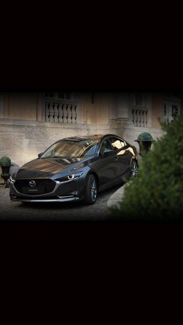 A black Madza3 sedan seen from the front parked next to a bush and an old stone house.