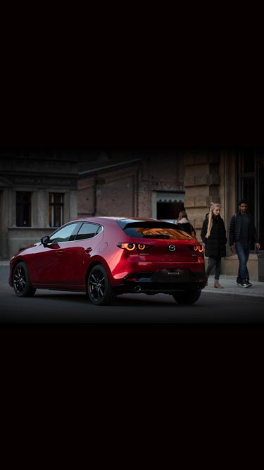 Vue arrière d’une Mazda3 Hatchback rouge, avec deux personnes à côté d’elle.