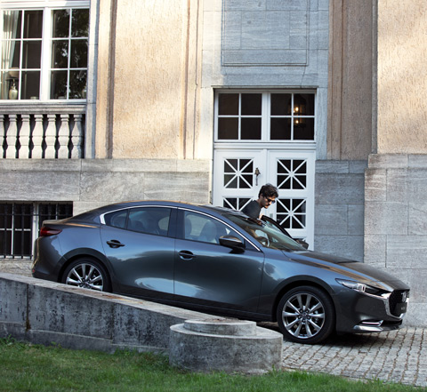 Una Mazda3 sedan grigia parcheggiata a lato di una strada in discesa con un uomo che le cammina accanto.