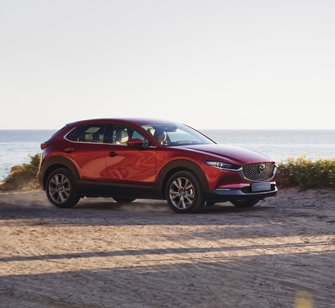 Potpuno nova crvena Mazda CX-30 snimljena s boka dok je parkirana pokraj plaže.