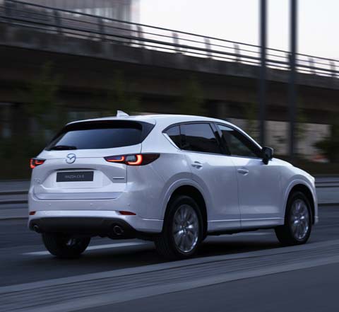 En hvid 2023 Mazda CX-5 kører ned ad en vej fotograferet bagfra.