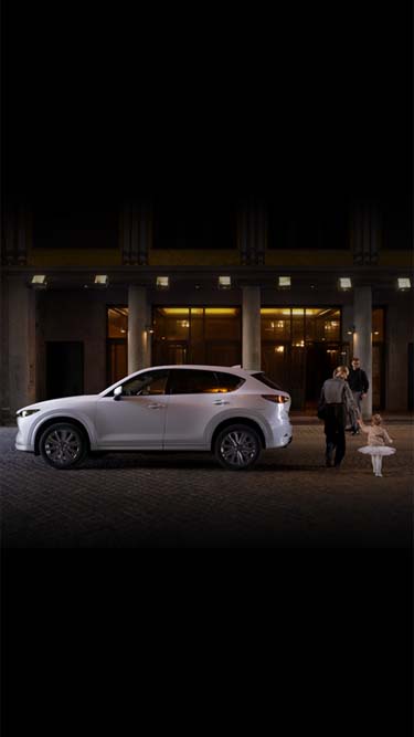 Par koji vodi dete na balet sa belim automobilom Mazda CX-5 parkiranim na ulici.