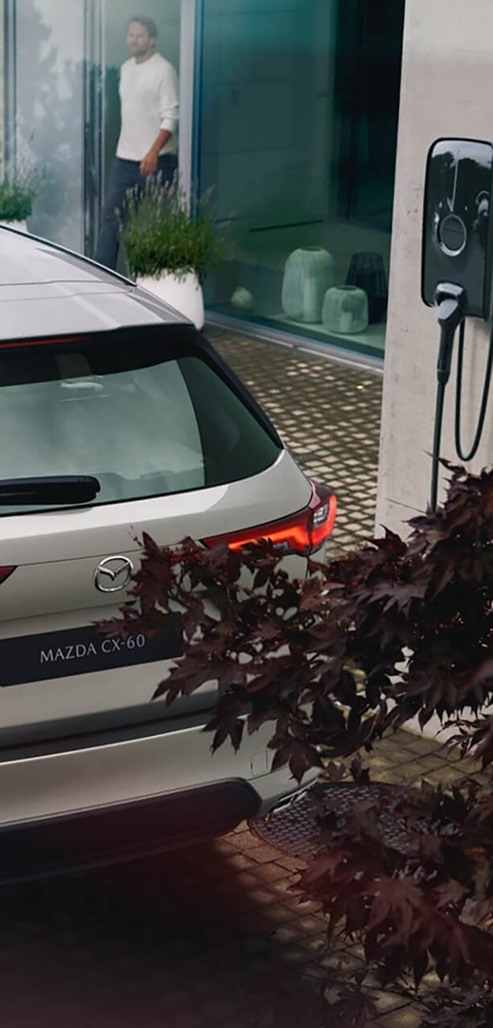Novi priključnohibridni SUV Mazda CX-60 ob zidni omarici za polnjenje doma.