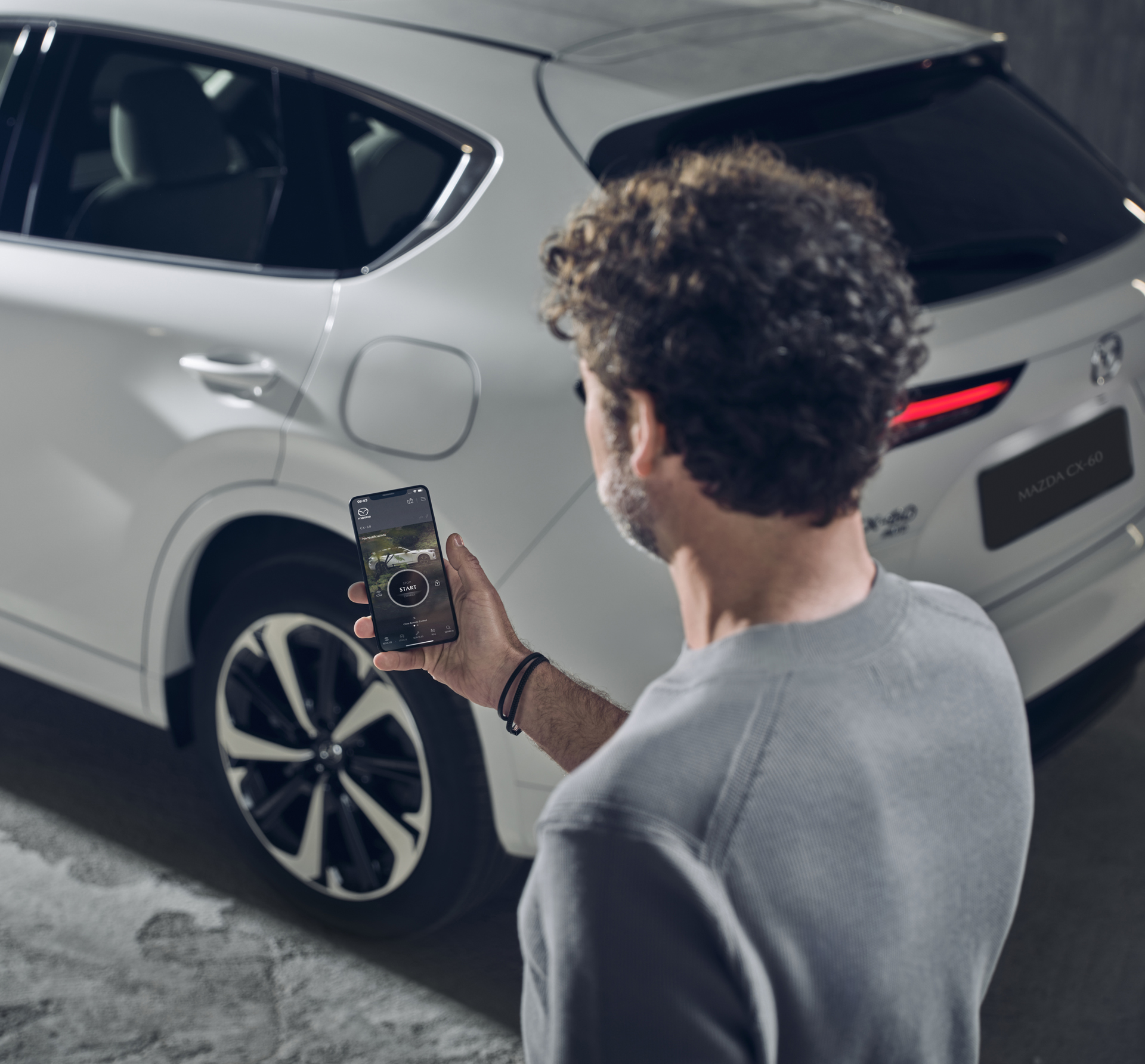 Мъж, използващ приложението MyMazda на смартфон, стоящ до бяла Mazda CX-60 Plug-In Hybrid.