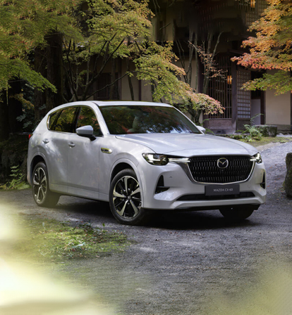 Den nye Mazda CX-60 Plug-In Hybrid SUV vist forfra parkeret foran en eng fyldt med træer.