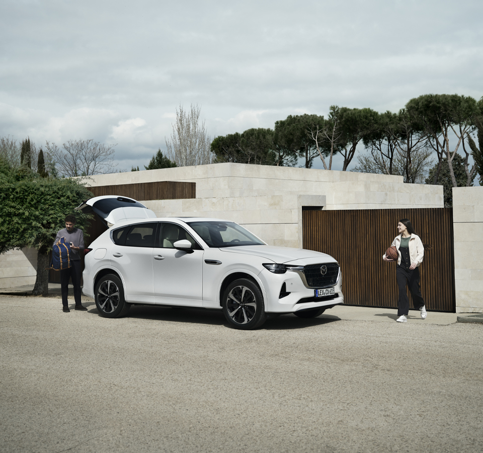 Un véhicule hybride rechargeable Mazda CX-60 blanc garé avec un homme et une fille se tenant debout à l’extérieur, un ballon de basketball à la main.