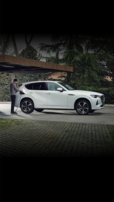 Balts uzlādējamais hibrīdautomobilis Mazda CX-60 novietots pie modernas mājas, ko ieskauj daudz koku.