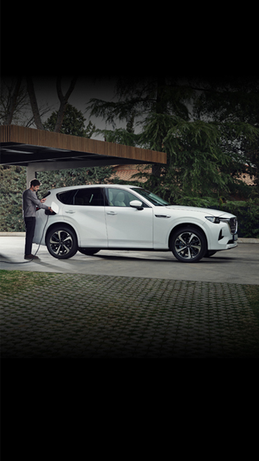 Biała Mazda CX-60 Plug-In Hybrid zaparkowana przed nowoczesnym domem otoczonym wieloma drzewami.