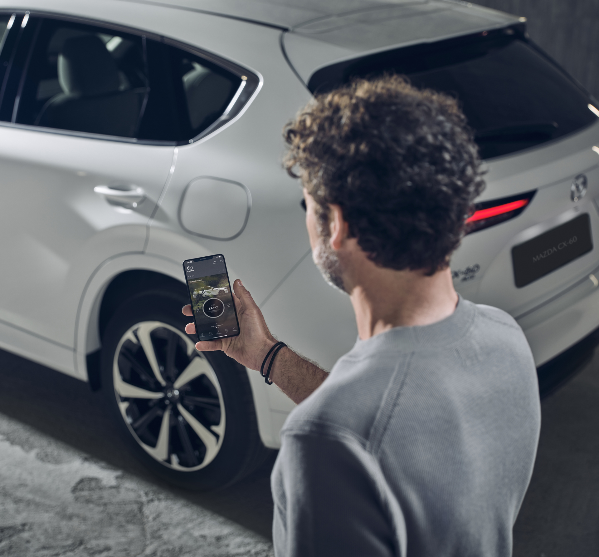 Ein Mann, der die MyMazda App auf einem Smartphone verwendet, steht neben einem weißen aufladbaren Hybrid-SUV Mazda CX-60.