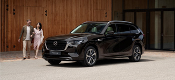 Mazda CX-80, паркирана пред сграда с мъж и жена, вървящи около нея.