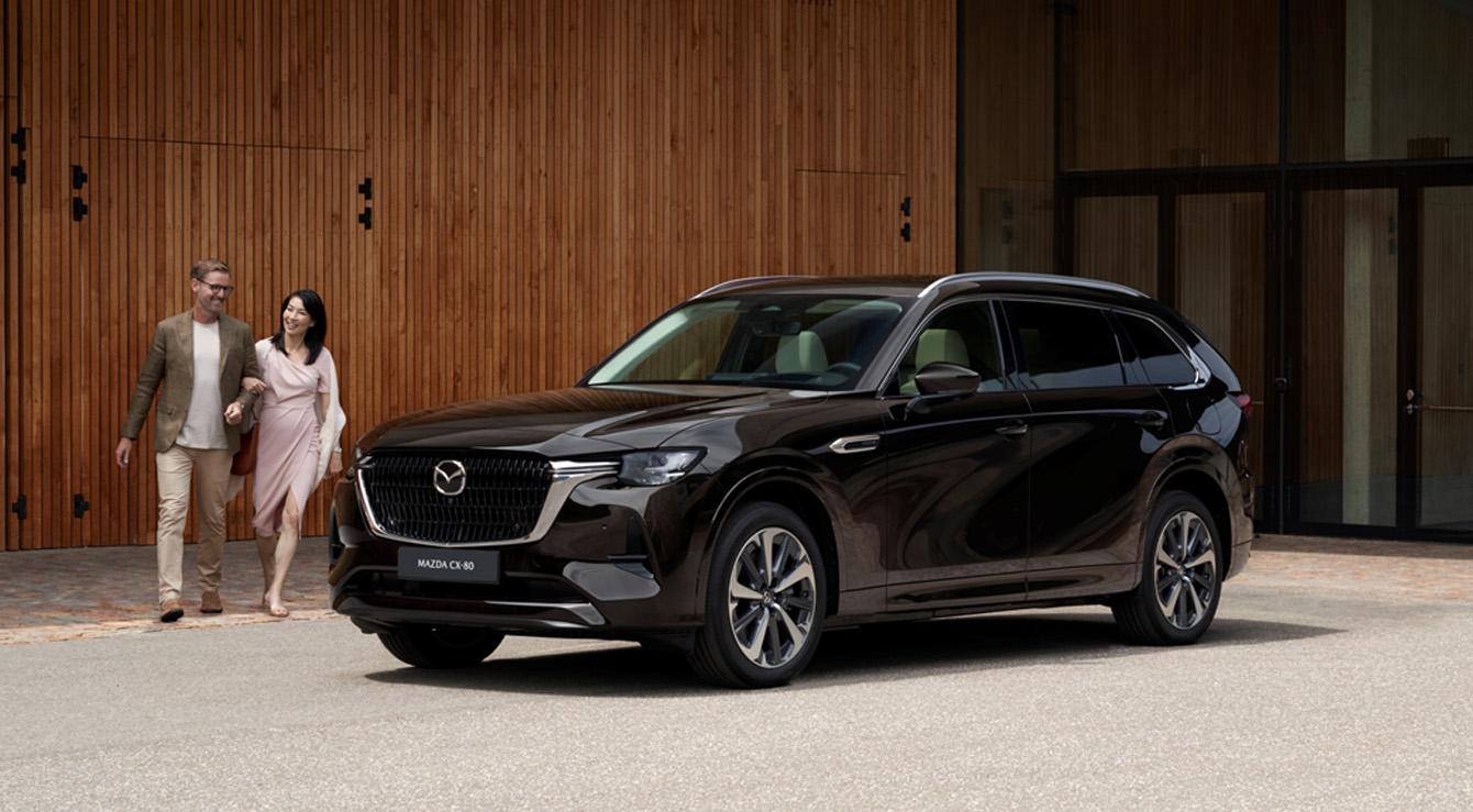 Černá Mazda CX-80 SUV zaparkovaná před zdí a pár, který kráčí kolem ní.