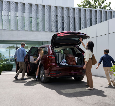 Rodina nastupuje do SUV Mazda CX-80 s pohonem všech kol v červené barvě. Matka zavírá velké zadní dveře.