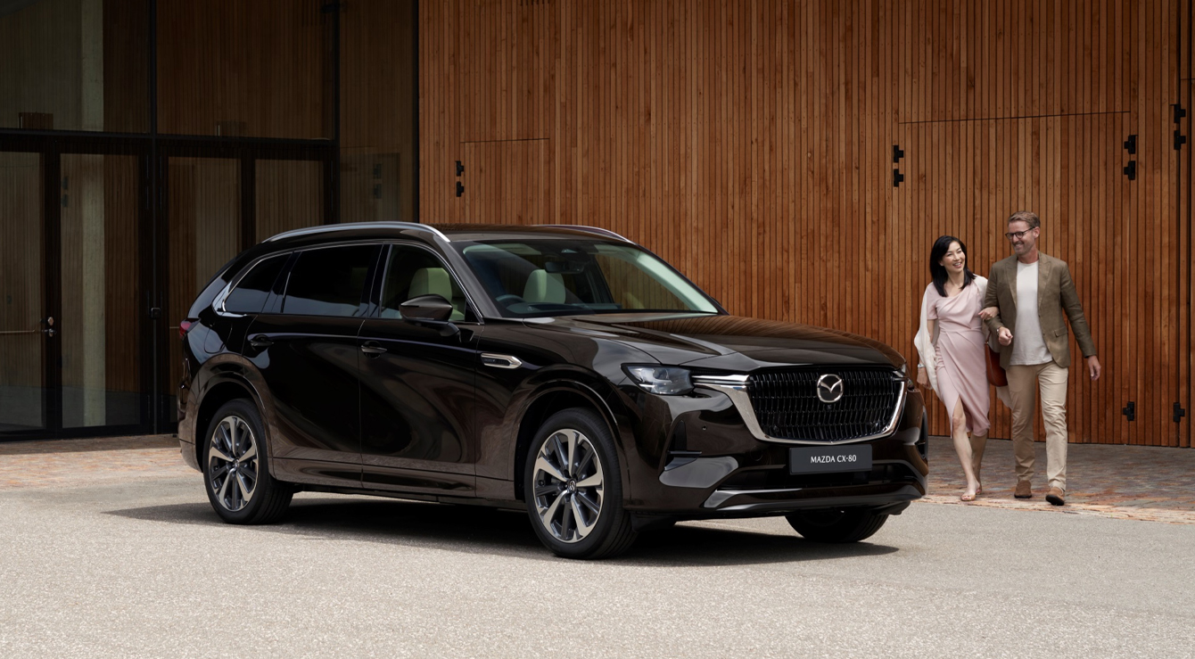 A black Mazda CX-80 SUV parked in front of a wall and a couple walking around it.