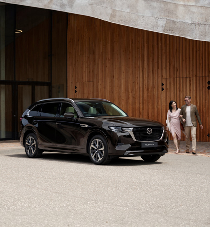 A black Mazda CX-80 SUV parked in front of a wall and a couple walking around it.