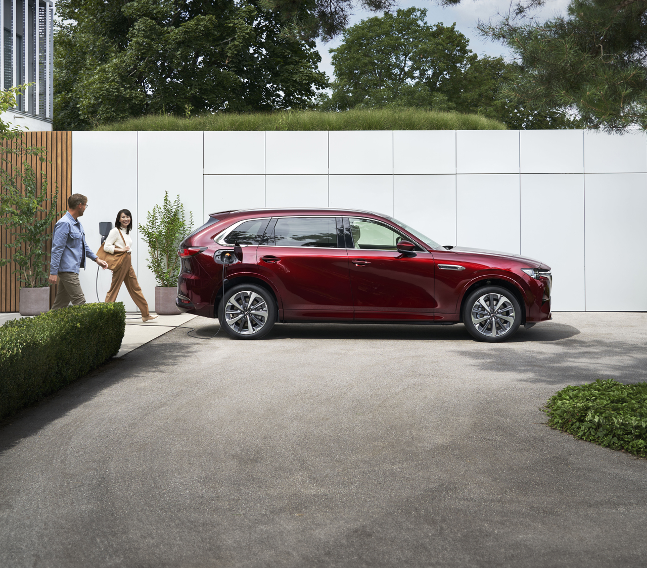 Une Mazda CX-80 rouge garée devant un mur et un couple marchant à côté de celle-ci.