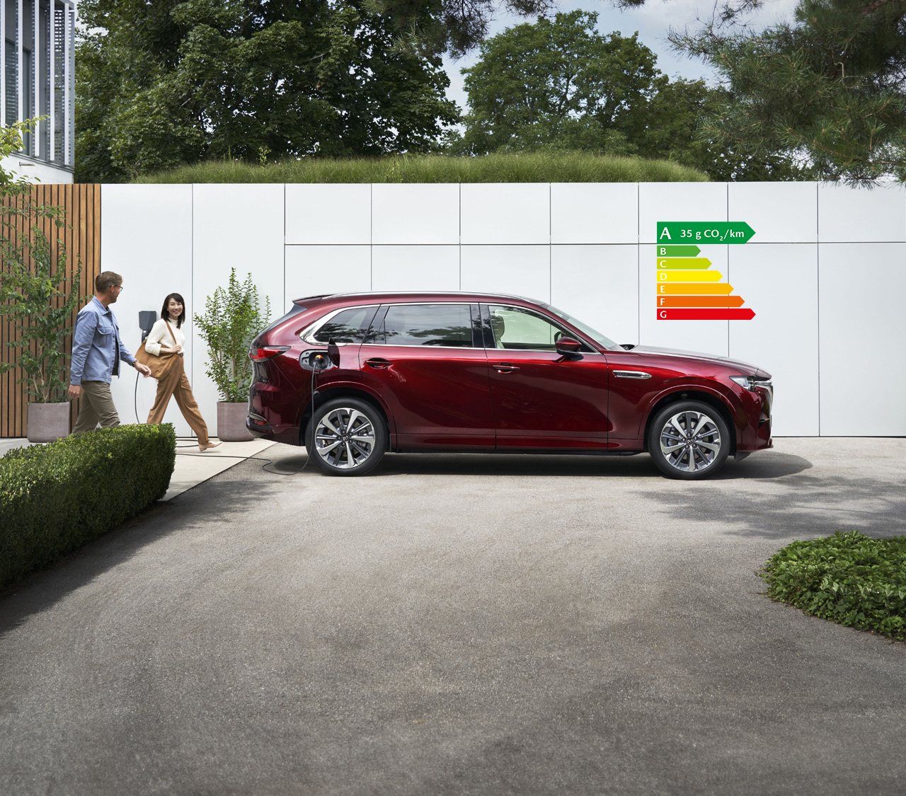 Mazda CX-80 rouge stationné devant un mur avec un couple marchant à côté.