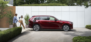 Mazda CX-80 zaparkowana przed budynkiem, za którą spaceruje para.