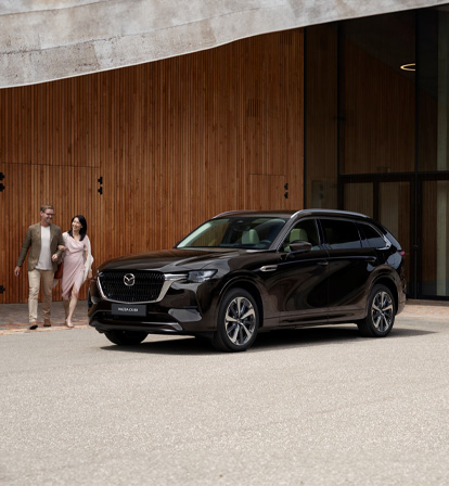 SUV Mazda CX-80 preto estacionado em frente a um muro e um casal a andar à sua volta.